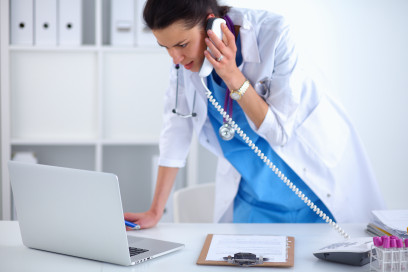 Young woman doctor in white coat at computer using phone